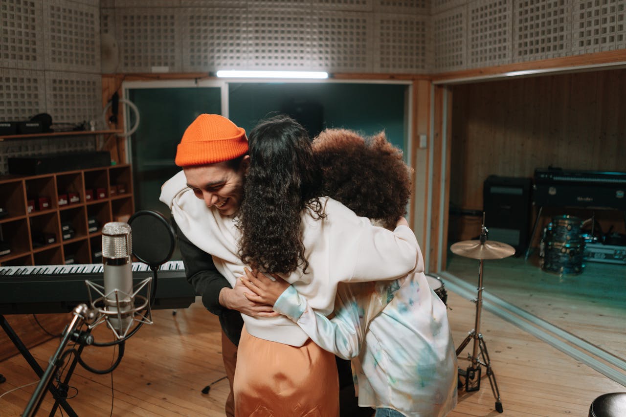 Three people warmly embrace in a music recording studio, showcasing friendship and teamwork.